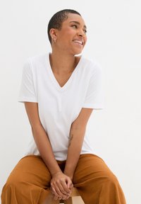 T-shirt bianca a v, realizzata in tessuto morbido, con maniche corte. Abbinata a pantaloni beige larghi, seduta su uno sgabello di legno.