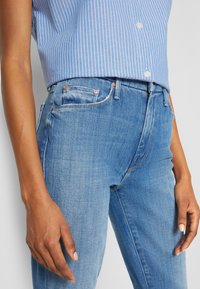 Persoon draagt een lichtblauw gestreept overhemd met knopen, ingestopt in een blauwe spijkerbroek met een normale taille, met ontspannen handen langs het lichaam.