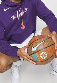 Sweat à capuche violet avec graphisme "el Valle" et numéro "1", associé à un short blanc. Tenant un ballon de basket Nike multicolore avec un logo floral.