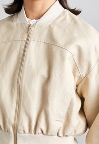 Beige bomberjack met een ritssluiting, ribgebreide kraag en manchetten, en een gerimpelde onderrand, gemaakt van zachte, gestructureerde stof.