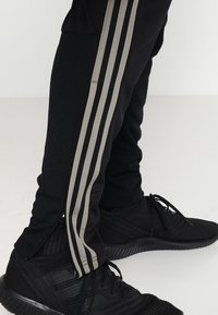Schwarze Sporthosen mit drei vertikalen grauen Streifen an den Seiten, mit Reißverschlussöffnungen an den Knöcheln und einer strukturierten Oberfläche. Dazu schwarze Sneakers.
