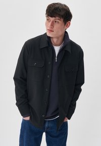 Fekete gombos ing-jacket két mellzsebbel, egy tengerészkék cipzáras pulóver fölött viselve, kék farmernadrággal párosítva. Simább anyag és strukturált fazon.