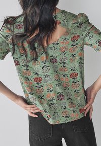 Blusa floral verde de mangas cortas, con escote en V y espalda descubierta. La tela tiene varios patrones de flores en rojo, naranja y púrpura.
