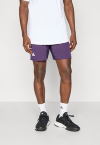 Shorts de sport violets en matériau léger, arborant un logo Adidas. Associés à un t-shirt blanc, des baskets noires et des chaussettes blanches.