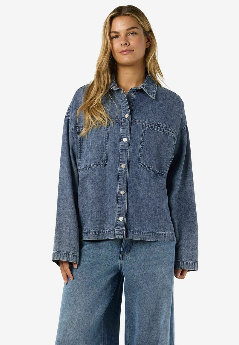 Chaqueta vaquera en azul oscuro con cuello, cierre de botones, dos bolsillos en el pecho y hombros caídos. Tejido delgado y texturizado con un ajuste holgado.