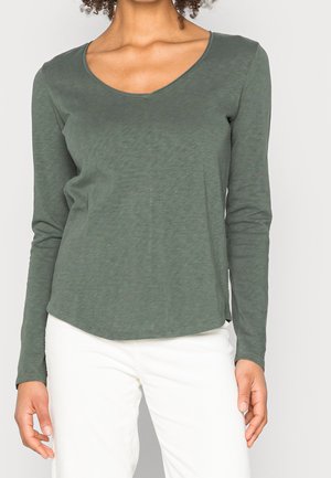 Frau trägt ein olivgrünes Langarmshirt mit V-Ausschnitt und weiße Hose, steht mit locker an den Seiten hängenden Händen vor einem schlichten Hintergrund.