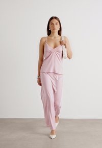 Lyserød satin cami med plisseret detalje, parret med matchende lette bukser. Modellen bærer en lys bæltepose og har hvide hæle på.