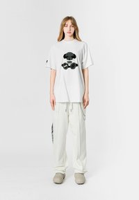 Camiseta blanca de gran tamaño con un diseño gráfico de un oso negro. Combinada con pantalones cargo blancos holgados y calzado texturizado en beige.