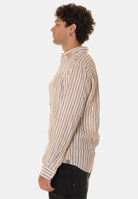 Camicia a maniche lunghe a righe nei toni beige, arancione e grigio; presenta un colletto con bottoni e una vestibilità su misura. Tessuto texturizzato con cuciture visibili.