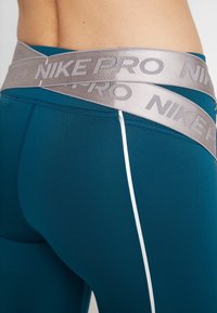 Turkusowe legginsy sportowe z grubym srebrnym pasem Nike Pro. Cechują się eleganckim wyglądem z białymi akcentami wzdłuż boków.