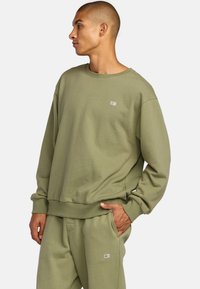 Olivgrüner sweatshirt aus weichem Stoff, mit Rundhalsausschnitt, gerippten Bündchen und einem kleinen Logopatch auf der Brust. Lockerer Schnitt.