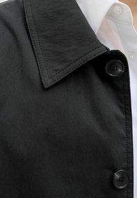 Jachetă din material textil negru, cu un guler mare, având o textură netedă, închidere cu nasturi negri și o cămașă albă dedesubt.