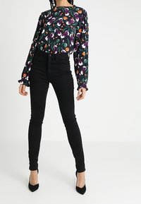 Sorte skinny jeans parret med en langærmet, mønstret bluse med multikolorede blomsterdesigns på en mørk baggrund.