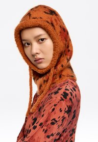 Capucha de punto con un patrón naranja y un acabado texturizado, que presenta lazos, combinada con una blusa de color óxido que muestra un patrón abstracto negro.
