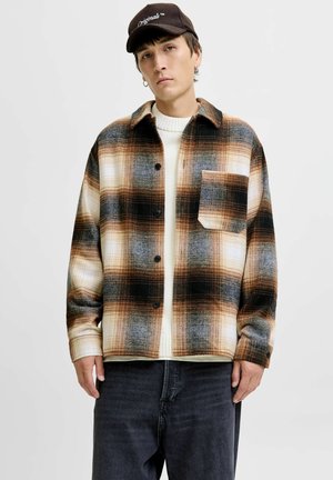 Flannel shirt in brown, black, and cream plaid pattern; button-up front; single chest pocket; worn over a white sweater with ribbed texture.