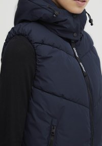 Marinblå vadderad väst med hög krage, med dragkedja, justerbar dragsko på huven och logotypdetalj på dragkedjan.