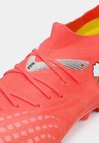 Zapatilla de fútbol de color rojo brillante con material de malla texturizada, cordones delgados y detalles plateados, con un patrón distintivo en la zona de los dedos.