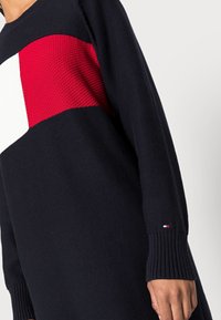 Pull marine avec un design horizontal audacieux en sections rouges et blanches. Tissu tricoté texturé, poignets côtelés et petit logo détaillé.