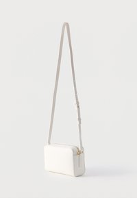 Μικρή λευκή ορθογώνια τσάντα χιαστί με ρυθμιζόμενο ιμάντα και χρυστό φερμουάρ σε απλό ανοιχτό φόντο.