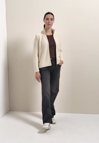 Femme aux cheveux foncés attachés en queue de cheval portant une polaire crème zippée, un haut marron, un pantalon noir et des baskets blanches, debout contre un mur beige.