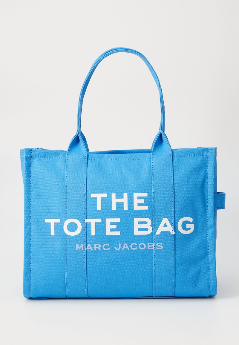 Saco de lona azul com alças duplas e texto branco "THE TOTE BAG MARC JACOBS" impresso na frente.