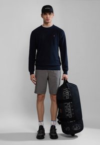 Marineblå trøje, grå shorts, sorte sneakers og en sort duffeltaske med hvidt logo-print. Model står mod en grå baggrund.