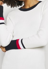 Frau trägt einen gerippten weißen Pullover mit schwarzem Kragen und Bündchen, der rote, weiße und schwarze Streifen aufweist, die Arme vor der Brust verschränkt.