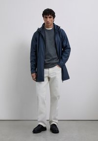 Hombre con una chaqueta de lluvia con capucha en color navy, suéter gris, jeans blancos y zapatos negros, de pie contra una pared clara y lisa.