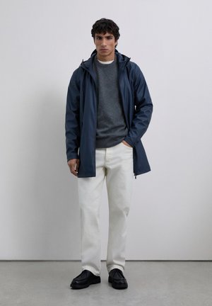 Mann in einer navyblauen, kapuzenbewehrten Regenjacke, grauem Pullover, weißen Jeans und schwarzen Schuhen, der vor einer schlichten hellen Wand steht.