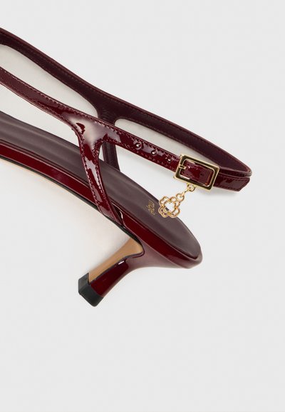 Burgundijas laku ādas papēžu sandeļi ar elegantu dizainu, regulējamu sprādzi un dekoratīvu zelta piekariņu uz potītes siksniņas.