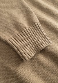 Pull beige en tricot avec un poignet côtelé. La texture est lisse avec un motif de points régulier, soulignant son aspect confortable.