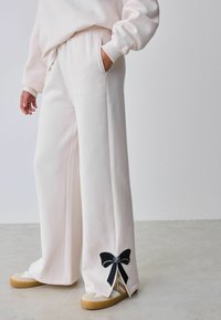 Cremefarbene Hose mit weitem Bein, Seitentaschen und schwarzer Schleifstickerei am Saum. Hergestellt aus weichem Stoff mit glatter Textur.