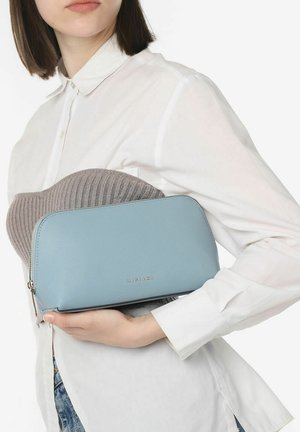 Bolso de mano texturizado en azul claro con herrajes plateados, que presenta un cierre de cremallera y un logotipo grabado, sostenido contra una camisa blanca de botones.