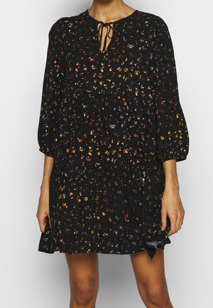Femme portant une robe noire à motifs floraux avec manches longues et un col noué, debout devant un fond uni.