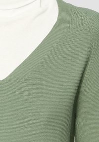 Groene gebreide trui met een V-halsontwerp, met een gestructureerde afwerking. Een witte coltrui is zichtbaar onder de trui.