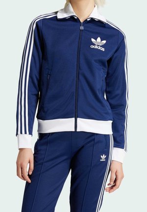 Personne portant un survêtement Adidas bleu marine avec des bandes blanches et un logo, composé d'une veste zippée et d'un pantalon assorti.
