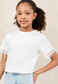 Hvid t-shirt med korte ærmer, der har detaljeret blondekant ved manchetterne, parret med lyseblå denimshorts og et enkelt armbånd.