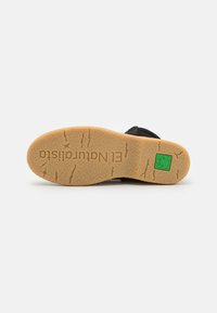 Suela de zapato de ante negro con una base de goma color caramelo, que presenta patrones texturizados y un acento de logo verde.