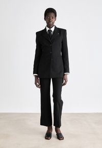 Blazer nero su misura con chiusura a un bottone, abbinato a pantaloni neri slim e mules neri con punta aperta. Camicia bianca a righe visibili.