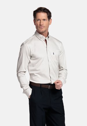 Mann trägt ein hellgraues Hemd mit Knöpfen, das in schwarze Hose mit braunem Gürtel gesteckt ist, steht mit einer Hand in der Tasche.