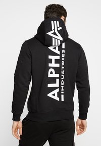 Felpa nera con grande scritta bianca "ALPHA INDUSTRIES" sul retro, dotata di cappuccio con coulisse e tasca con zip sulla manica.