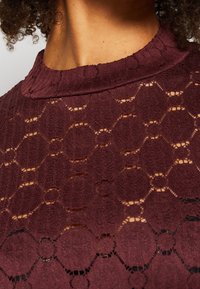 Burgundy stickad plagg med en strukturerad, honungscomb-mönster och en hög halsringning, med öppna stickade sektioner som skapar en lager-effekt.