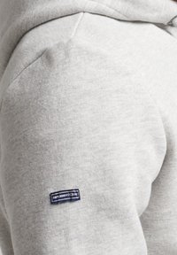 Sudadera gris claro con una textura suave, que presenta un parche de logo azul y blanco en el hombro izquierdo, y capucha.