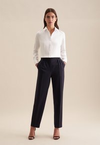 Witte blouse met kraag en lange mouwen, gecombineerd met marineblauwe plooibroek. De broek heeft een getailleerde pasvorm en zakken; gladde stofstructuur.