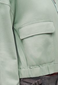 Κοντινό πλάνο του μανικιού ενός ανοιχτοπράσινου μπουφάν με μεγάλη τσέπη με καπάκι και ελαστική ρίγα στο τελείωμα, φορεμένο πάνω από σκούρο γκρι παντελόνι με ζώνη.