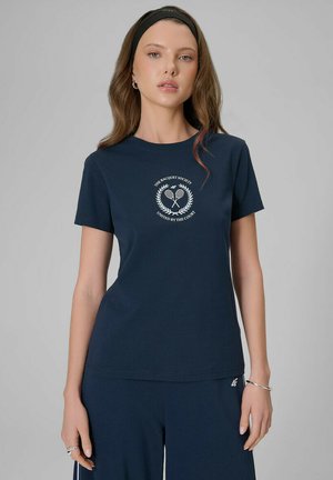 Kobieta o długich włosach, ubrana w granatowy T-shirt z logo rakiet tenisowych i dopasowane spodnie, uzupełniona opaską na głowę i srebrną bransoletką.