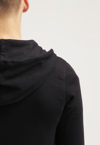 Schwarzer Hoodie mit einer strukturierten Kapuze, glattem Stoff und sichtbaren Nähdetails. Die Rückansicht zeigt eine lockere Passform und ein minimalistisches Design.