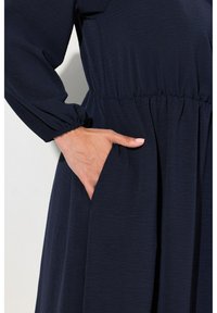 Navyblaues Kleid mit langen Ärmeln, elastischem Taillenbund und seitlicher Tasche, aus strukturiertem Stoff mit sanftem Fall.