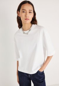 Balta pārliela izmēra T-krekls ar apaļu apkakli un uzlocītām piedurknēm, izgatavots no mīksta kokvilnas. Kā aksesuārs ir sudraba ķēdes kaklarota.