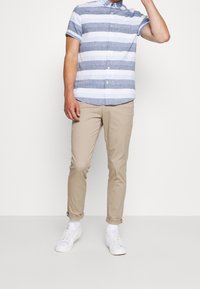 Blå och vitrandig kortärmad skjorta med knappar, ljusbeiga smala byxor och vita sneakers, stående mot en neutral bakgrund.